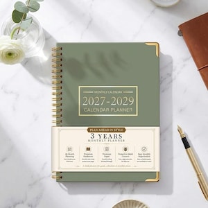 May include: A sage green spiral-bound monthly calendar planner for 2027-2029 with gold-tone metal corner protectors. A white paper band wraps around the center, featuring the text 3 YEARS MONTHLY PLANNER and listing features like premium hardcover and paper.