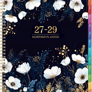 May include: A spiral-bound monthly planner with a dark navy blue cover featuring white flowers and gold foliage accents. The text 27-29 MONTHLY PLANNER is printed in gold in the center. The right edge has colorful monthly tabs from Jan to Dec.