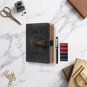 May include: A black journal with a sun and moon phase embossed cover, secured by a bronze-colored combination lock. The journal features copper-toned page edges and is accompanied by a black fountain pen and five small color swatches.