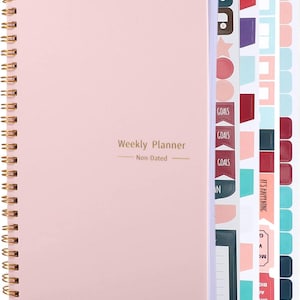 May include: A light pink spiral-bound weekly planner with gold-colored wire binding. The cover features the text "Weekly Planner" and "Non-Dated" in gold lettering. Included are several sheets of colorful adhesive stickers with various shapes.