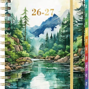 May include: A spiral-bound planner featuring a watercolor landscape of a river, forest, and mountains with the text 26-27 in gold. The planner includes a gold elastic closure and multi-colored monthly tabs along the right edge for organization.