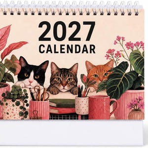May include: A 2027 desk calendar featuring an illustration of three cats peeking out from behind various potted plants, books, a pencil holder, and a pink mug. The background is a soft cream color with the text 2027 CALENDAR printed in black.
