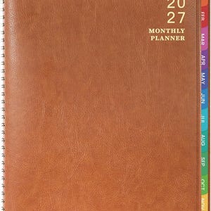 May include: A brown, textured, spiral-bound monthly planner for the year 2027. The cover features the gold-stamped text 20 27 MONTHLY PLANNER and a small crown icon. Multi-colored monthly index tabs are visible along the right edge of the planner.