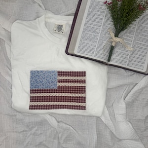 May include: White t-shirt with a sewn-on American flag design. The flag features a blue floral print field, red and white checkered stripes, and white lace trim. The shirt is displayed on a white, textured surface.