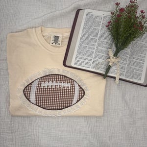 以下が含まれることがあります： クリーム色のクルーネックTシャツ。中央には茶色と白のギンガムチェック柄のフットボールのグラフィックが描かれ、白いステッチのレースと白いレースの縁取りが施されています。テキストが印刷された開いた本と、小さな赤い花のブーケが添えられています。