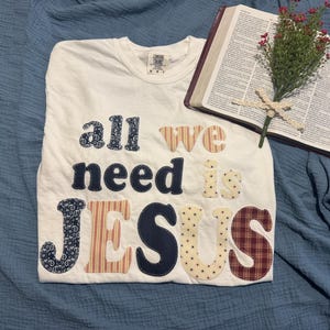 Puede incluir: Camiseta blanca con la frase "all we need is JESUS" en varias telas estampadas. Una Biblia abierta con un arreglo floral y una cruz se coloca junto a la camiseta sobre un fondo de tela azul.