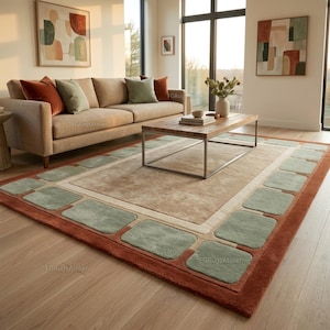 Può includere: Un tappeto rettangolare caratterizzato da un campo centrale beige, un bordo color crema e una cornice esterna di blocchi rettangolari verde salvia su un bordo color terracotta intenso. Il tappeto ha una consistenza morbida e un motivo geometrico.