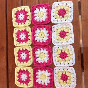 Peut inclure: Une collection de douze carrés de granny crochetés de différentes couleurs. Les carrés présentent un motif floral central rose, blanc et jaune, entouré d'une bordure contrastante. Les carrés sont disposés sur une surface en bois.