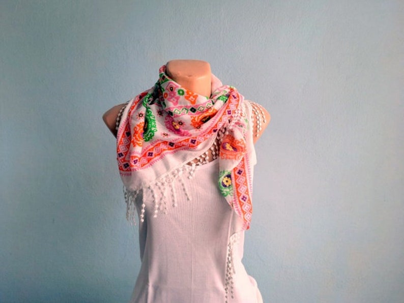 K&ouml;nnte beinhalten: Ein wei&szlig;er Schal mit einem rosa, orange und gr&uuml;nen Blumenmuster. Der Schal hat einen Fransenrand und ist um ein wei&szlig;es Tanktop gebunden.