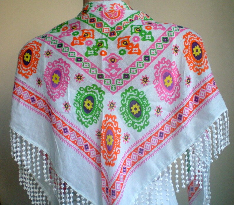 K&ouml;nnte beinhalten: Wei&szlig;er Baumwollschal mit einem farbenfrohen geometrischen Muster in den Farben Rosa, Orange, Gr&uuml;n und Lila. Der Schal hat einen Fransenrand.