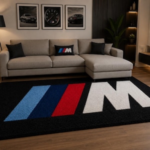 Op de afbeelding: Een rechthoekig zwart vloerkleed met het BMW M-logo, bestaande uit drie schuine strepen in lichtblauw, donkerblauw en rood, gevolgd door een grote witte letter M. Het kleed heeft een getextureerd, hoogpolig oppervlak en ligt op een vloer.