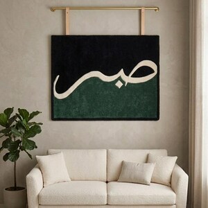 May include: A rectangular wall hanging featuring a tufted texture with a black upper section and a dark green lower section. The Arabic word Sabr is written in large, cream-colored script across the center. It is suspended from a gold metal rod.