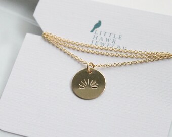 May include: A gold-colored necklace with a round pendant featuring a sunburst design. The necklace is displayed on a white card with the text "LITTLE HAWK JEWELRY" and a bird logo. The chain is delicate and the pendant is centered.