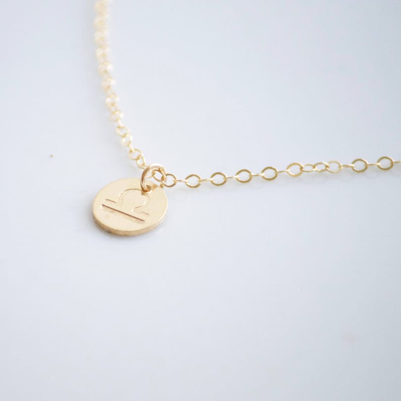 May include: Gold necklace with a Libra zodiac sign pendant. The pendant is a small, round disc with the Libra symbol engraved on it.