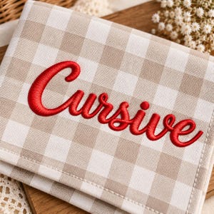 Puede incluir: Un artículo de tela rectangular con un patrón de cuadros vichy en beige y blanco. La palabra Cursive está bordada con hilo rojo intenso en el centro de la tela en una fuente cursiva. El artículo tiene costuras visibles a lo largo del borde inferior.