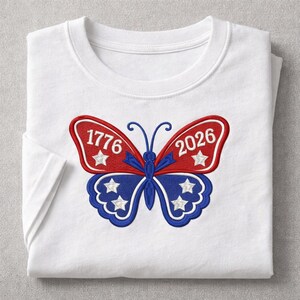 May include: White t-shirt with a patriotic butterfly design. The butterfly has red and blue wings with the numbers "1776" and "2026" and white stars. The shirt is folded.