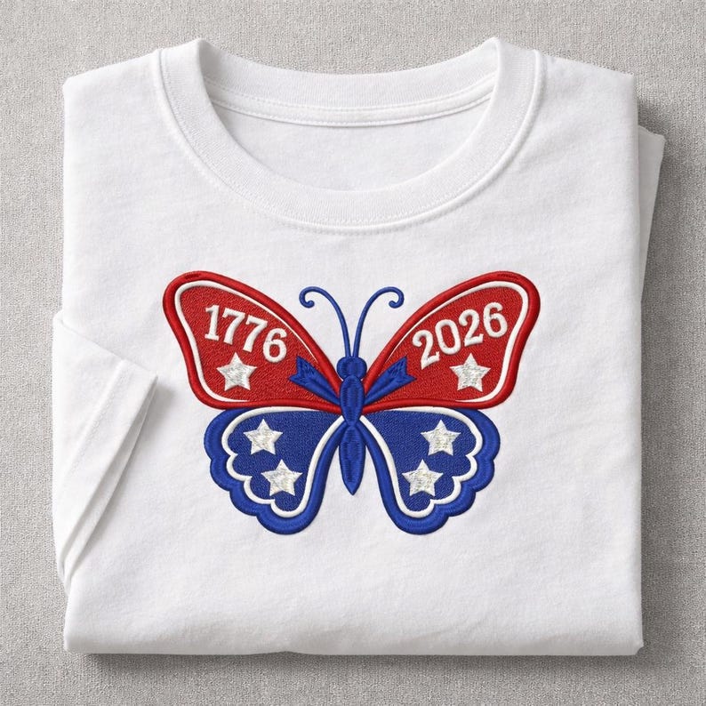 May include: White t-shirt with a patriotic butterfly design. The butterfly has red and blue wings with the numbers "1776" and "2026" and white stars. The shirt is folded.
