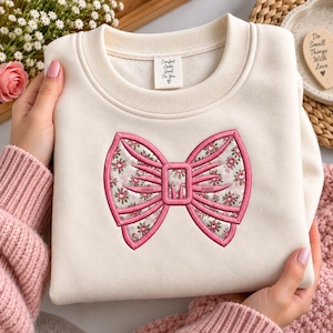 Peut inclure: Un sweat-shirt à col rond de couleur crème avec un grand nœud brodé sur le devant. Le nœud présente un contour cousu rose et un intérieur à motif floral. Une étiquette de col blanche affiche le texte Comfort Looks Good On You.