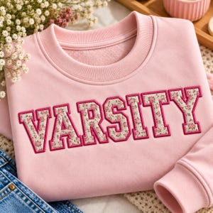 May include: A light pink crewneck sweatshirt featuring the word VARSITY in large block letters. The letters are filled with a cream-colored fabric patterned with small pink and green floral designs and outlined in a dark pink stitched border.
