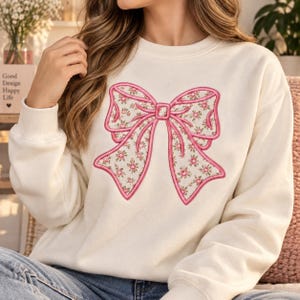 May include: A cream-coloured crewneck sweatshirt featuring a large embroidered bow on the chest. The bow has a pink stitched outline and is filled with a repeating pattern of small pink roses and green leaves on a white background.