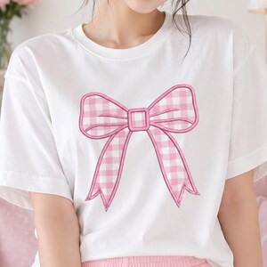 May include: White t-shirt featuring a large pink bow with a gingham pattern. The bow is outlined in a darker pink, creating a contrast against the white fabric. The shirt has short sleeves and a round neckline.