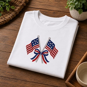 Op de afbeelding: Wit T-shirt met ronde hals met een geborduurd ontwerp van twee gekruiste Amerikaanse vlaggen met een rood, wit en blauw lint. De vlaggen hebben rood-witte strepen en een blauw veld met witte sterren. Het shirt is opgevouwen.