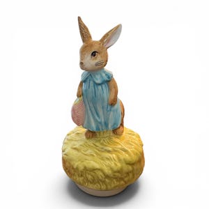 Puede incluir: Figura de cerámica de un conejo marrón con un vestido azul y una cesta rosa. El conejo está sobre una base de paja amarilla. La figura tiene detalles finos y está sobre un fondo blanco.