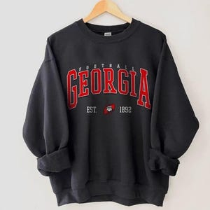 May include: Black sweatshirt featuring the word "GEORGIA" in red block letters with a white outline. The word "FOOTBALL" is above the word "GEORGIA". Below the word "GEORGIA" is the text "EST. 1892" with a bulldog logo.