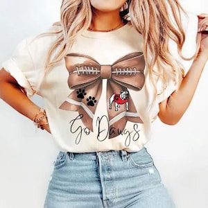 May include: Cream-coloured t-shirt with a brown American football-shaped bow design, paw prints, and a bulldog in a red sweater. The words "Go Dawgs" are written in a script font below the bow.