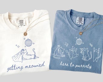 Op de afbeelding: Twee T-shirts, één witte en één blauwe, met kat-thema graphics. Het witte shirt zegt "getting meowied" met een kat met sluier. Het blauwe shirt zegt "here to purrrty" met feestende katten. Beide shirts hebben een ronde gouden hangerketting.