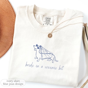 May include: An ivory short-sleeve t-shirt featuring a blue line-art graphic of a dachshund wearing sunglasses and a bridal veil. The text below the dog reads bride in a weenie bit. The shirt has a Comfort Colors label indicating size S.