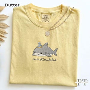 May include: A butter yellow short-sleeve t-shirt featuring an embroidered gray shark wearing a blue party hat with yellow polka dots. Below the shark, the word overstimulated is embroidered in black. A silver chain necklace rests on the collar.