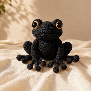 Peut inclure: Une grenouille en crochet noir dotée de grands yeux ronds aux iris jaunes marqués et aux pupilles noires. La grenouille présente une surface texturée en fil, des membres définis avec des doigts distincts et un corps arrondi en position assise.