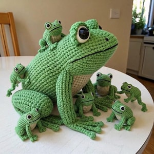 Könnte beinhalten: Ein großer gehäkelter grüner Frosch mit cremefarbenem Bauch und großen schwarzen Augen, begleitet von sieben kleineren gehäkelten grünen Fröschen in verschiedenen Größen. Die Frösche haben eine strukturierte Maschenoptik auf weißer Fläche.