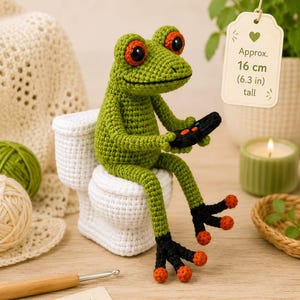 Könnte beinhalten: Ein grüner gehäkelter Frosch mit großen orangefarbenen Augen sitzt auf einer weißen gehäkelten Toilette und hält einen schwarzen Gamecontroller. Ein Etikett am Frosch gibt eine Höhe von ca. 16 cm an. Der Frosch hat schwarze Füße mit orangefarbenen Zehen.
