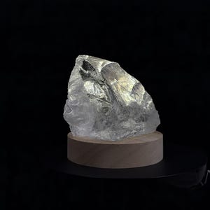 Pode incluir: Uma lâmpada de rocha cristalina transparente e irregular assenta sobre uma base circular de madeira clara. A pedra translúcida apresenta arestas e facetas afiadas e irregulares que captam a luz, criando um efeito brilhante e luminoso contra o fundo escuro.