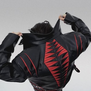 Puede incluir: Chaqueta de cuero negra con un llamativo diseño geométrico rojo en la espalda. La chaqueta tiene un cuello alto y detalles en rojo. La espalda presenta un patrón rojo y negro audaz. La chaqueta se abrocha con una hebilla.
