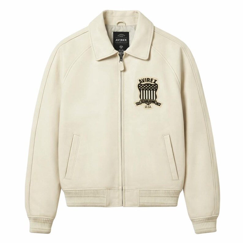 May include: Cream-colored leather jacket with a zip-up front and a collared neckline. Features a black and white emblem on the chest with the text "AVIREX" and "U.S.A.". Ribbed cuffs and hem complete the design.