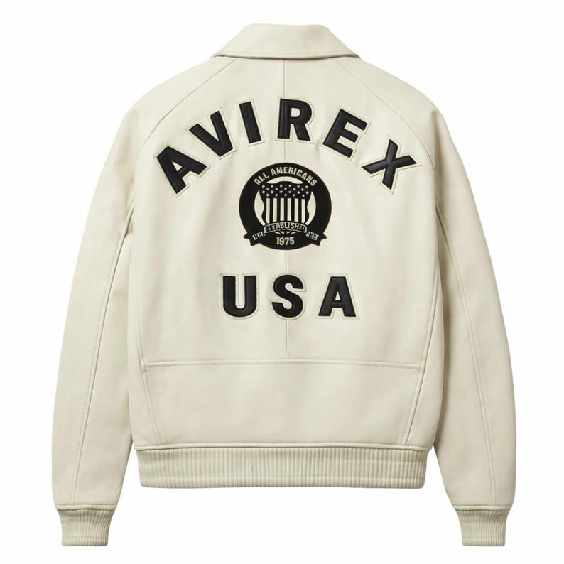 May include: Cream-colored leather jacket with a classic collar and ribbed cuffs. The back features the word "AVIREX" arched across the top, with a circular emblem in the center and "USA" below. The emblem includes the text "ALL AMERICANS ESTABLISHED 1975".