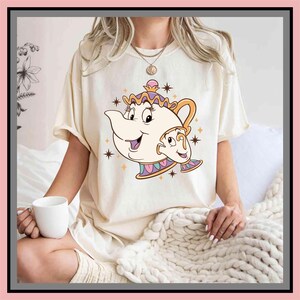 Può includere: T-shirt color crema con un'illustrazione a cartoni animati di Mrs. Bric e Chicco de La Bella e la Bestia. Il design include una teiera e una tazza sorridenti con accenti dorati, rosa e viola. La maglietta ha una vestibilità ampia.