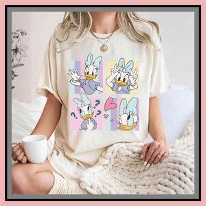 May include: Cream-colored t-shirt featuring a Daisy Duck graphic. The design is divided into four squares, each showcasing a different pose of Daisy Duck. The squares have pastel backgrounds of pink, blue, and yellow. The shirt has short sleeves.