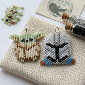 May include: Two beaded pendants, one depicting a character with large ears in green, tan, and brown, and the other a helmet design in gray, black, and blue. The pendants are on a beige fabric with small glass bead accents and two small glass bottles.