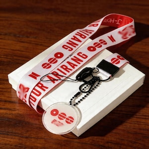 May include: A white lanyard with red text and graphics, including the words "TOUR ARIRANG" and "J-HOPE." The lanyard is draped over a white box, with a black clasp and a clear, circular charm with red graphics.