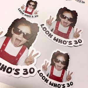 Może przedstawiać: Zestaw naklejek z dwoma r&oacute;żnymi wzorami. Jeden przedstawia małe dziecko w musztardowej koszuli z napisem "LOOK WHO'S 40". Drugi przedstawia dziecko w okularach przeciwsłonecznych i czerwonym kombinezonie z napisem "LOOK WHO'S 30".