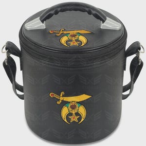 May include: A black cylindrical bag with a top handle and side strap attachments. The bag features embroidered gold scimitar and crescent moon emblems on the lid and front, accented with red and teal details. The surface has a repeating logo pattern.