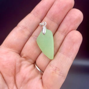 May include: A pale lime green sea glass pendant with a smooth, matte, frosted texture. The triangular-shaped piece of glass is securely attached to a polished silver-toned metal bail at the top, designed for threading onto a necklace chain.