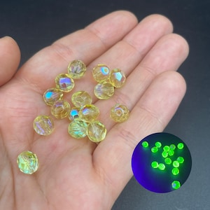 May include: A collection of small, round, faceted yellow beads with an iridescent finish. An inset circular graphic shows the same beads glowing with a bright neon green light when exposed to ultraviolet light, highlighting their fluorescent properties.