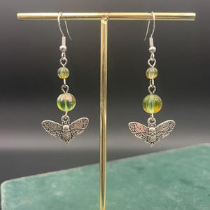 May include: A pair of silver-toned earrings featuring a detailed bee charm. Each earring has a series of green and yellow beads, suspended from a silver hook. The earrings are displayed on a gold-colored stand against a dark background.