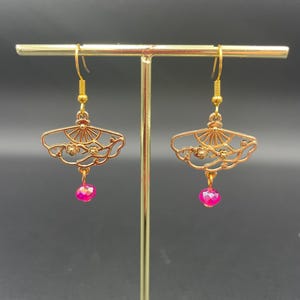 May include: Gold-coloured earrings with a fan-shaped design, intricate cutouts, and floral details. Each earring has a dangling, faceted, hot pink bead. The earrings are displayed on a gold-coloured jewellery stand against a dark background.