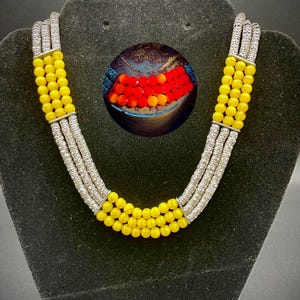May include: A multi-strand necklace with yellow beads and silver-colored accents. The necklace is displayed on a black velvet jewelry stand. A circular image of red and orange beads is in the background.